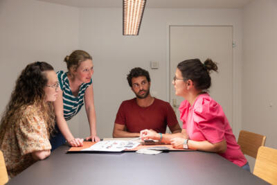 Vier Teammitglieder von Studio C sitzen (eine Person steht) um den Besprechungtisch des Büros, vor ihnen liegen Materialmuster und Pläne