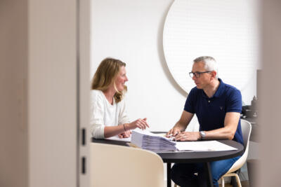 Markus Berweger und Mareike Buch sitzen im Büro von STudio C an einem Tisch vor Plänen, Mareike Buch spricht und deutet auf einen Plan